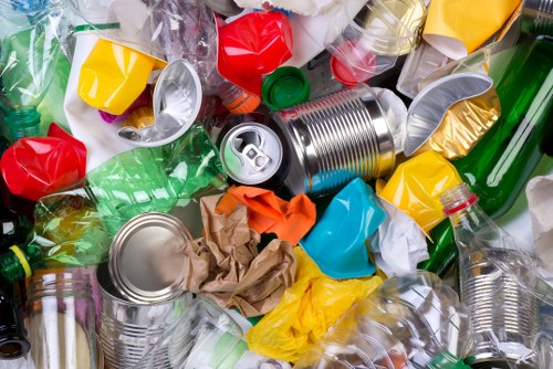A close-up view of a large pile of mixed recyclable and waste materials including crushed aluminum cans with silver surfaces, crumpled and torn paper in shades of brown, yellow, and orange, and several plastic bottles in green, clear, and red with some lids attached, scattered on a paved surface likely outdoors. The background shows additional discarded plastics and metals, suggesting a collection of rubbish prime for removal services. This scene reflects typical rubbish clutter encountered in waste removal projects around Barking, with various textures and colors highlighting the diverse types of waste managed by commercial waste removal specialists such as Commercial Waste Removal Barking. The overall composition emphasizes the need for effective rubbish collection and disposal, capturing a realistic snapshot of waste accumulation requiring professional clearance in the local area.