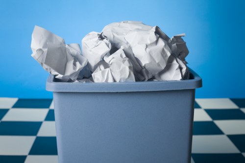 A blue plastic rubbish bin filled with crumpled white paper and paper waste, situated on a checkered black and white tiled floor. The bin is positioned against a smooth, light blue wall, with the paper waste overflowing slightly at the top, indicating recent filling or removal. The crumpled paper varies in size and texture, some with visible creases and folds, and the bin appears to be standard in size and finish, typical of waste disposal containers used in commercial or domestic settings. The environment suggests an indoor area, such as a kitchen, office, or utility space, aligned with the services provided by Commercial Waste Removal Barking for waste collection and management in the local postcode area. The lighting is even and bright, emphasizing the textures and the arrangement of the waste within the bin, highlighting the importance of professional rubbish removal in maintaining clean and organised premises.