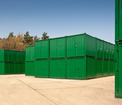 A large outdoor storage area featuring multiple green metal shipping containers stacked and arranged in a neat formation, situated on a paved surface. The containers have vertical ridges and are painted in a vivid green colour with some small vents and locking mechanisms visible on their front doors. The setting appears to be an industrial or commercial site, possibly a waste management or rubbish disposal facility, with a backdrop of trees and a clear blue sky. The containers are positioned to maximize space, with some stacked two high on the right and others aligned side by side, creating a compact storage configuration. The environment is well-lit, suggesting daytime, and the scene is free of debris or other visible objects, emphasizing the orderly nature typical of waste collection and disposal sites managed by companies like Commercial Waste Removal Barking, serving the local Barking area and postcode region.