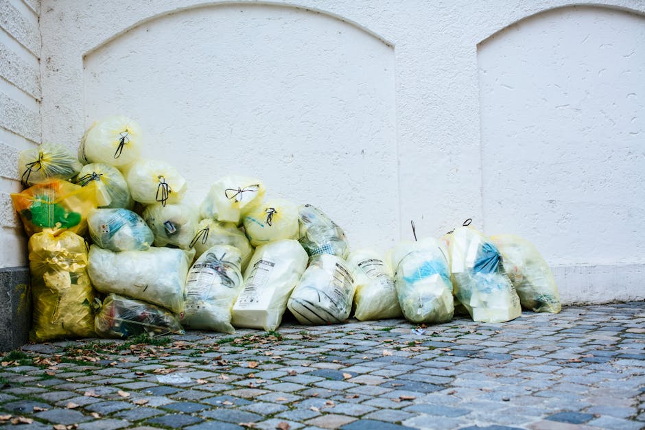 A collection of numerous plastic rubbish bags filled with waste, leaning against a white exterior wall on a cobblestone pavement. The bags are in various shades of yellow, white, and transparent, and