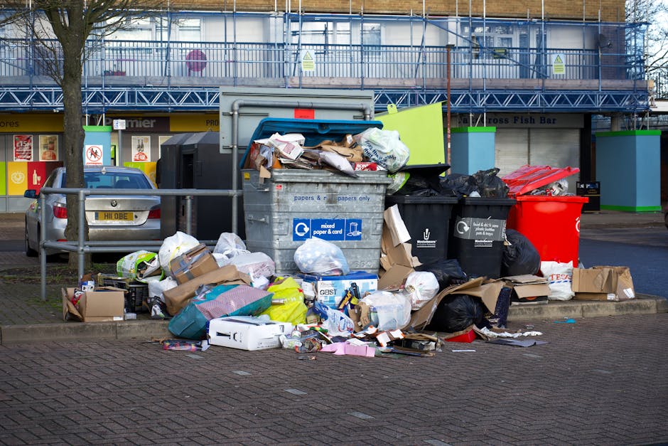 A sanitation worker in an orange high-visibility vest is performing waste collection or cleaning activity at the rear of a large red garbage truck parked on a roadside. The truck is equipped with meta