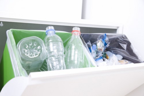 A close-up view of a waste collection bin with a bright green interior and a white exterior, situated inside a clean, modern environment. The bin contains clear, recyclable plastic bottles, one with a blue cap and the other with a reddish cap, both standing upright and partially visible. Next to these bottles, there is a black plastic bag filled with various mixed waste materials, including more plastic packaging. The surrounding area appears to be a well-lit space, possibly part of a commercial or residential premises in Barking, with the main focus on the sorted waste intended for removal service. The image reflects typical rubbish disposal activities relevant to commercial waste removal services in the local area, emphasizing plastic bottle recycling and general waste segregation.